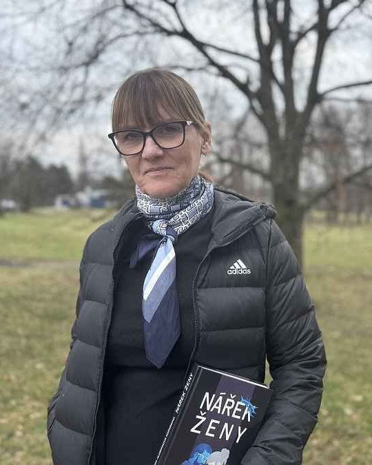 Ivana H. Kozmin outdoors in a park, holding her novel Nářek ženy