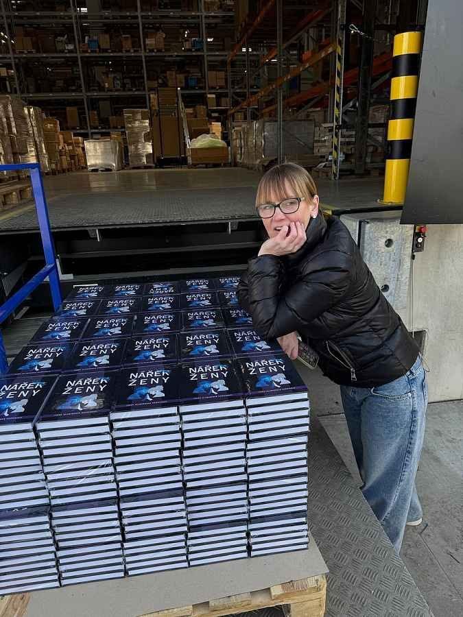 Ivana leaning on a pallet stacked with copies of Nářek ženy in a warehouse