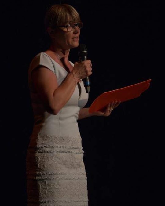 Ivana on stage with a microphone and a red folder